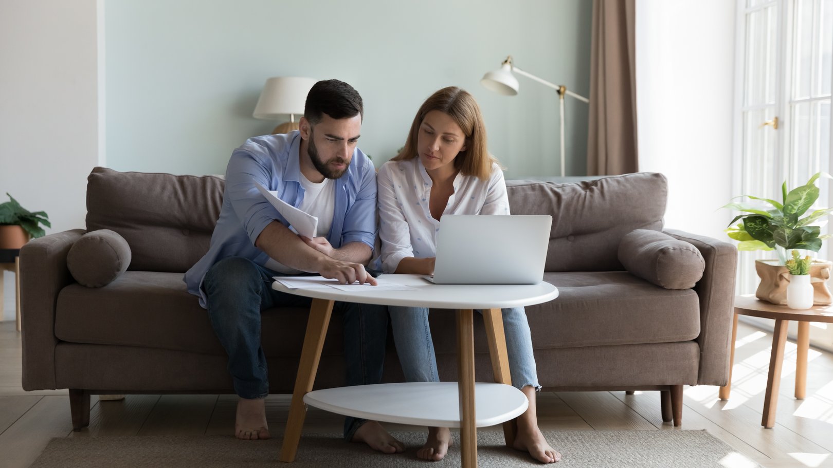 Serious millennial couple sit on sofa sorting out papers and payments on laptop